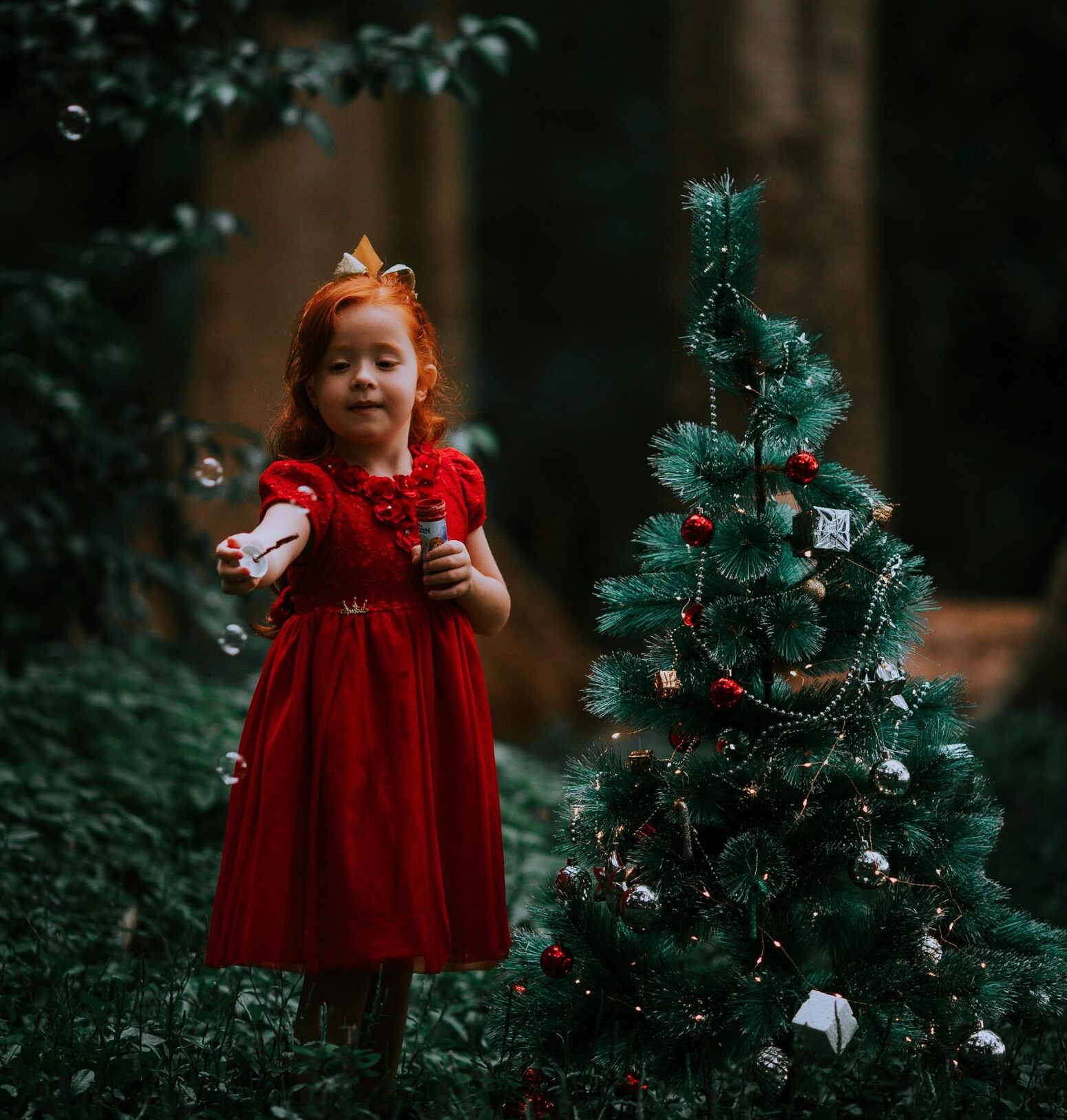 bambina in un bosco che coglie un fiore accanto a un albero di Natale, atmosfera fiabesca invernale